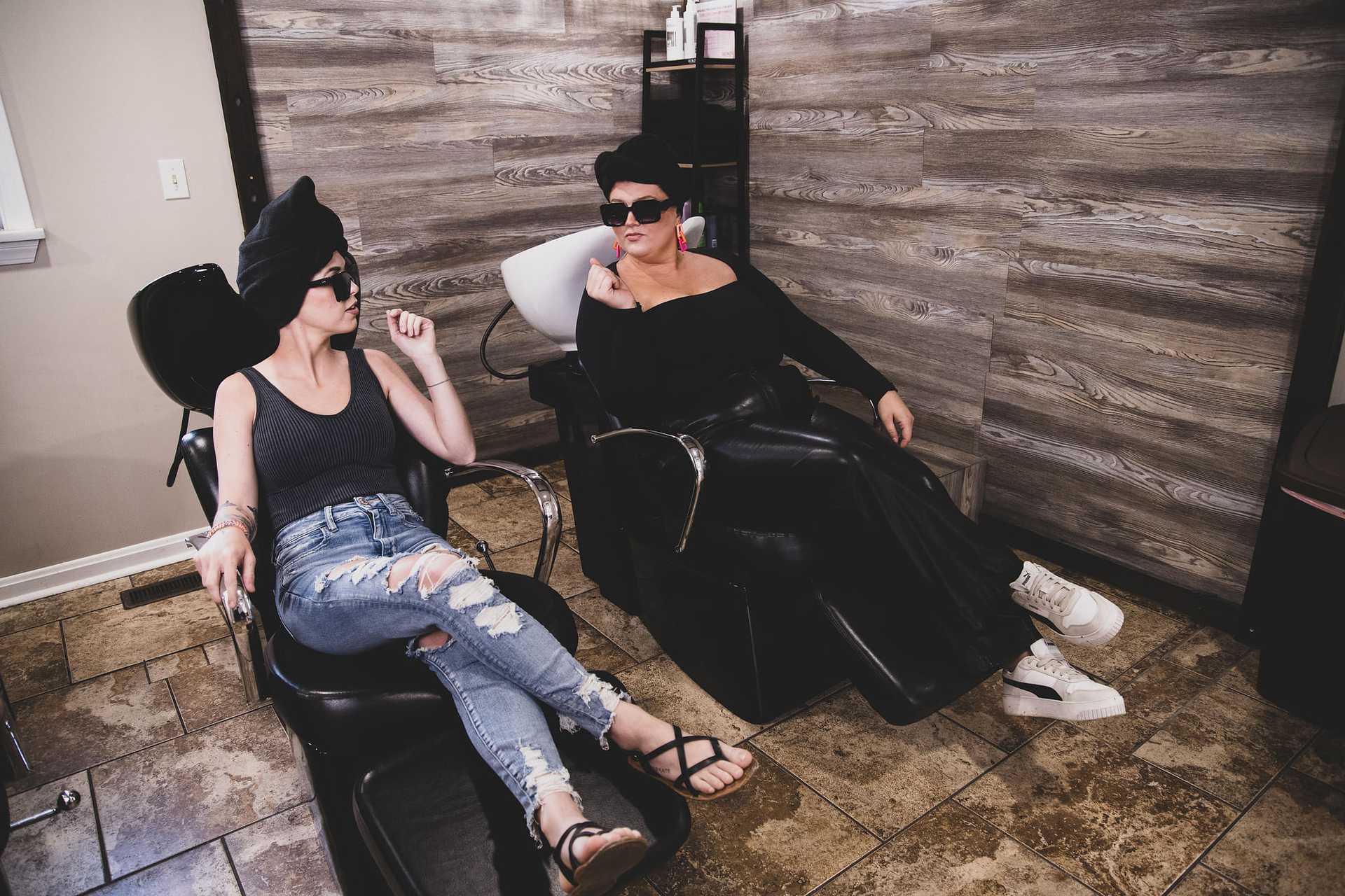 Two women relax in salon chairs, chatting and wearing black outfits and sunglasses.